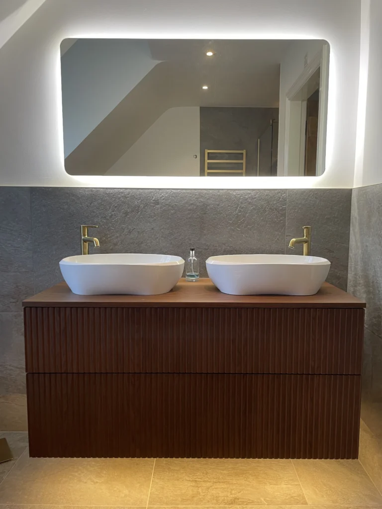 A modern bathroom vanity with a ribbed wood-effect cabinet, two white oval countertop basins with gold-colored faucets, a clear glass bottle between the sinks, a large rectangular mirror with a built-in LED light strip along the top, and light beige floor tiles.
